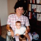 Photo: The image shows Laurie and her two children in her living room. The mother, Laurie, is sitting in her power wheelchair in the center of the photo, smiling, holding her six-month-old infant daughter in her lap with her 5 1/2-year-old son, also smiling, sitting on the foot pedals with his feet resting on the floor, leaning back against her legs. All of them are facing forward.  This photo was take in 1986.