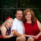 Photo: Image shows the Kidd family, family of three.  Picture is taken outside at their home. The young daughter is sitting to the left, wearing a dress with a red bow in her hair.  In the middle is the dad, wearing a polo shirt and shorts.  To the right is the mom, wearing a red dress.  They are sitting on a red outdoor loveseat, facing forward.