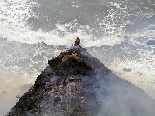 Los Padres firefighter Kevin Poyner (facing camera) and John Knight of the Big Sur Volunteer Fire Brigade appear to have one foot “in the black” and the other in the Pacific Ocean. In reality, the two men have ample room between their position and the cliffs leading down to the beach.