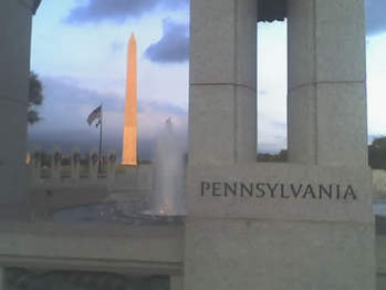 The Pennsylvania pillar at the World War II Monument stands in the foreground as the sun reflects on the American flag and Washington Monument.