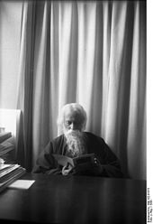 An old bearded man garbed in a dark mantle is reading from a slim book perched in his hands. He is sitting at a dark-toned desk cleared of everything but a neat stack of papers at left; in the background is a light-coloured curtain.