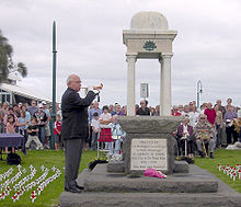 Photo of an ANZAC memorial with an elderly man playing a bugle. Rows of people are seated behind the memorial. Many small white crosses with red poppies have been stuck into the lawn in rows on either side of the memorial.