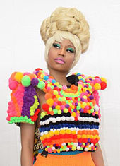 An Afro-American woman in a blonde wig and bright teal eyeshadow wears a shirt constructed of variously colored cotton balls.