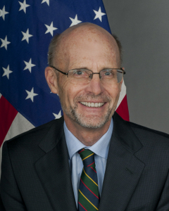 Man sitting in front of an American flag. (Photo Credit: State Department)