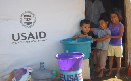 Children gather to have their photo taken in front of the temporary shelter built by USAID and implementing partner, Save the Children, in Patulúl, Suchitepéquez. The shelters were built as part of the humanitarian assistance response after Tropical Storm Stan destroyed villages and crop lands for hundreds of thousands of families in the Western Highlands and coastal areas of Guatemala in 2005. In the foreground is a can of cooking oil that is one of the food rations included in the food distribution programs to help families get back on their feet after losing everything but their lives. Photo Credit: Sonia Dominguez, USAID, 2006.