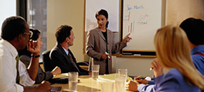a woman standing at a whiteboard lecturing other people around a table