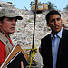 USAID Administrator speaks with a man wearing a USAID hat and vest, with rubble in the background