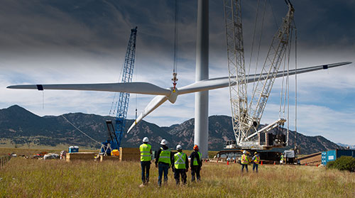 Photo of construction workers watching a crane lift the blades of a large wind turbine that is being constructed.