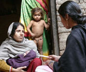 Woman holds swaddled baby in her arms, listening to woman across from her who has back to camera, another young child looks on