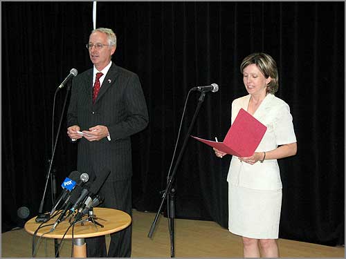 A man speaking at a microphone as a woman reads a document