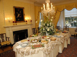 Dining room, lit by chandelier, with table filled with serving dishes of food, fireplace in the background