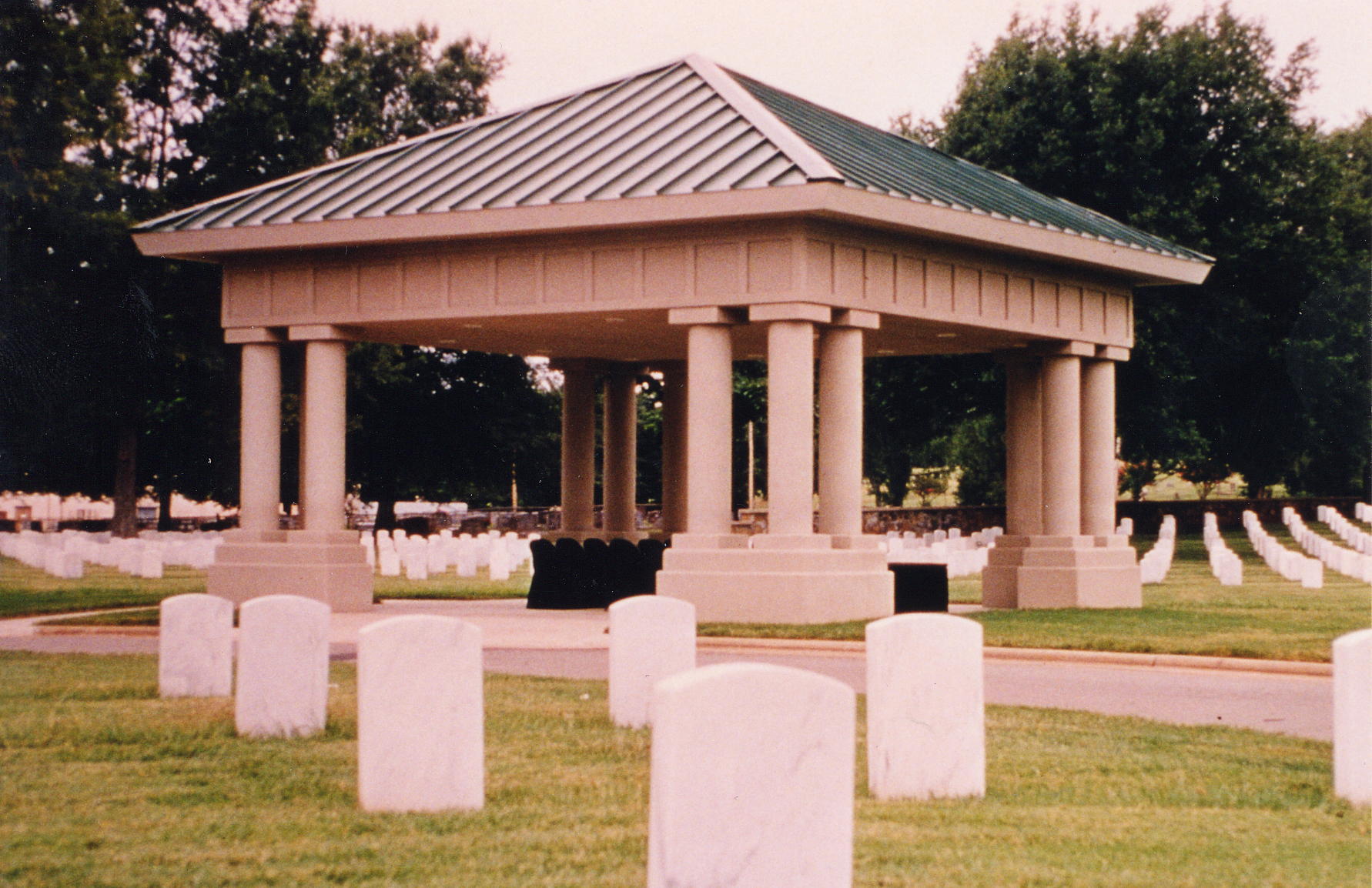 A photo of an open committal shelter rectangular in shape with three stone columns at each corner. Upright markers surround the shelter on three sides.