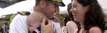 A male Service Member hugging his baby while smiling at his wife.