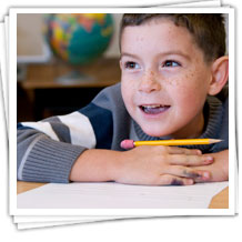 Boy leaning forward on his desk, hands resting on top of papers and a pencil balanced on his hands.