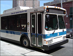 Photo of the front and part of the side of a bus parked at the curb of a city street with tall buildings in the background.