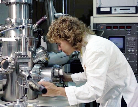 Photograph of a young woman in a white lab coat working with a piece of machinery.