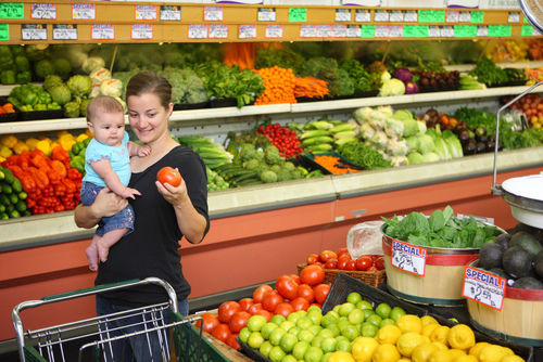 Young mother with baby selects items in produce aisle of grocery store.