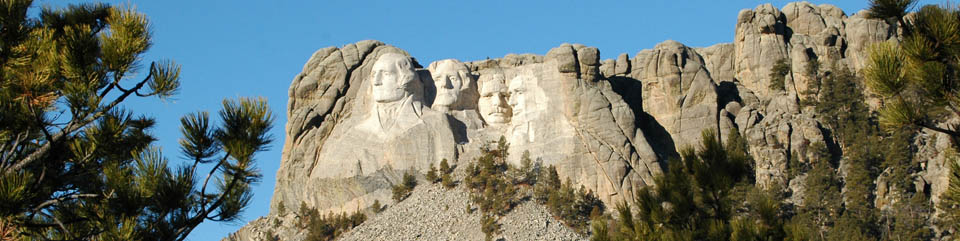 Mount Rushmore, Washington, Jefferson, T. Roosevelt, Lincoln framed by ponderosa pine trees under a bright blue sky.