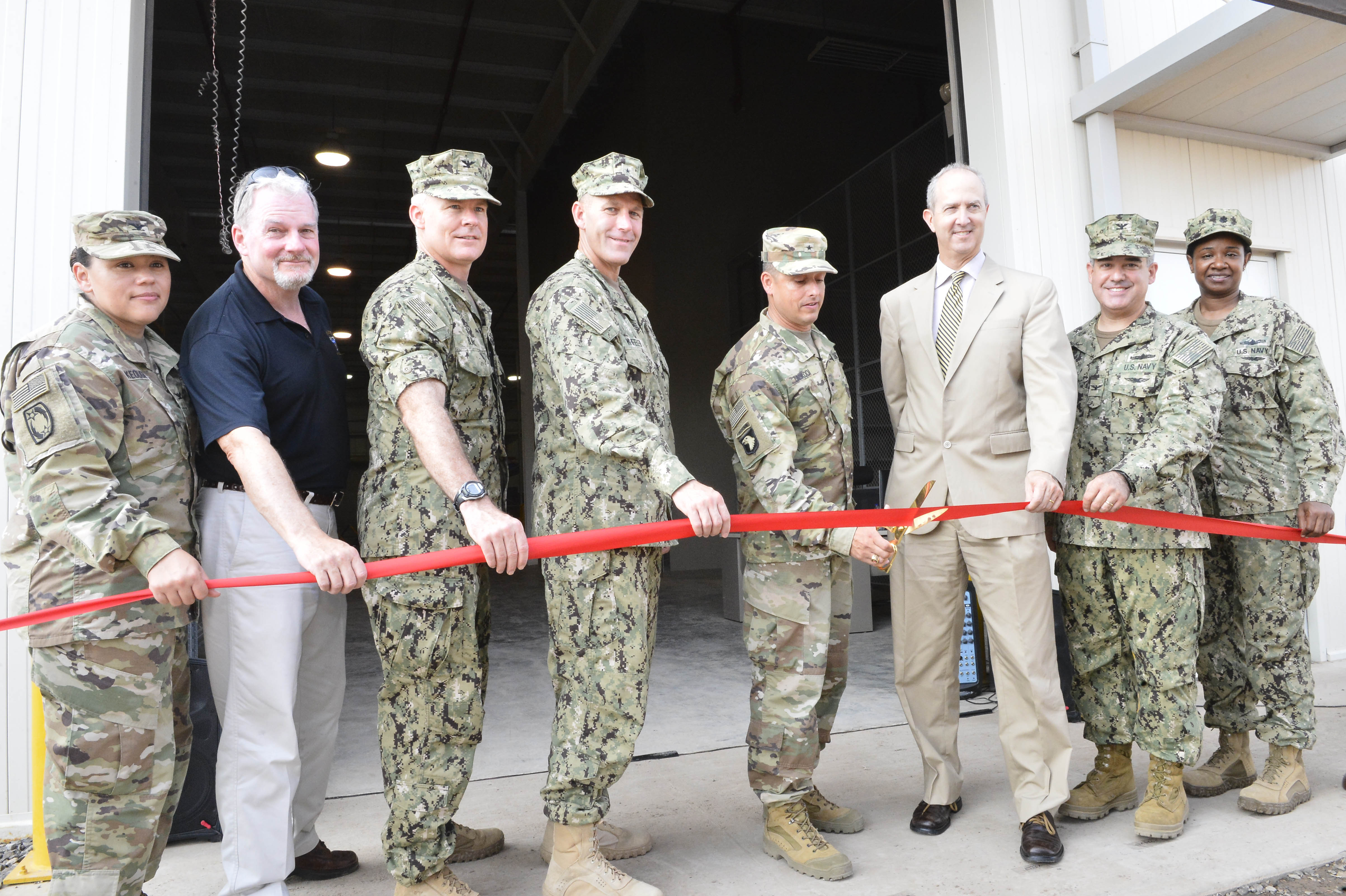 Facing viewer, six military personnel in fatigues, two civilians prepare to cut red ribbon