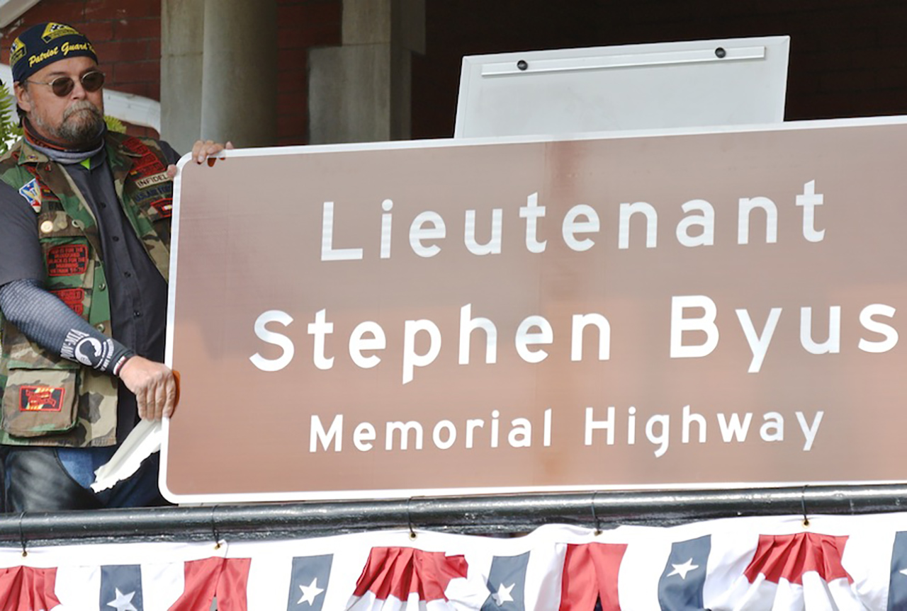Highway sign reading 'Lieutenant Stephen Byus Memorial Highway' held by man at left in biker apparel