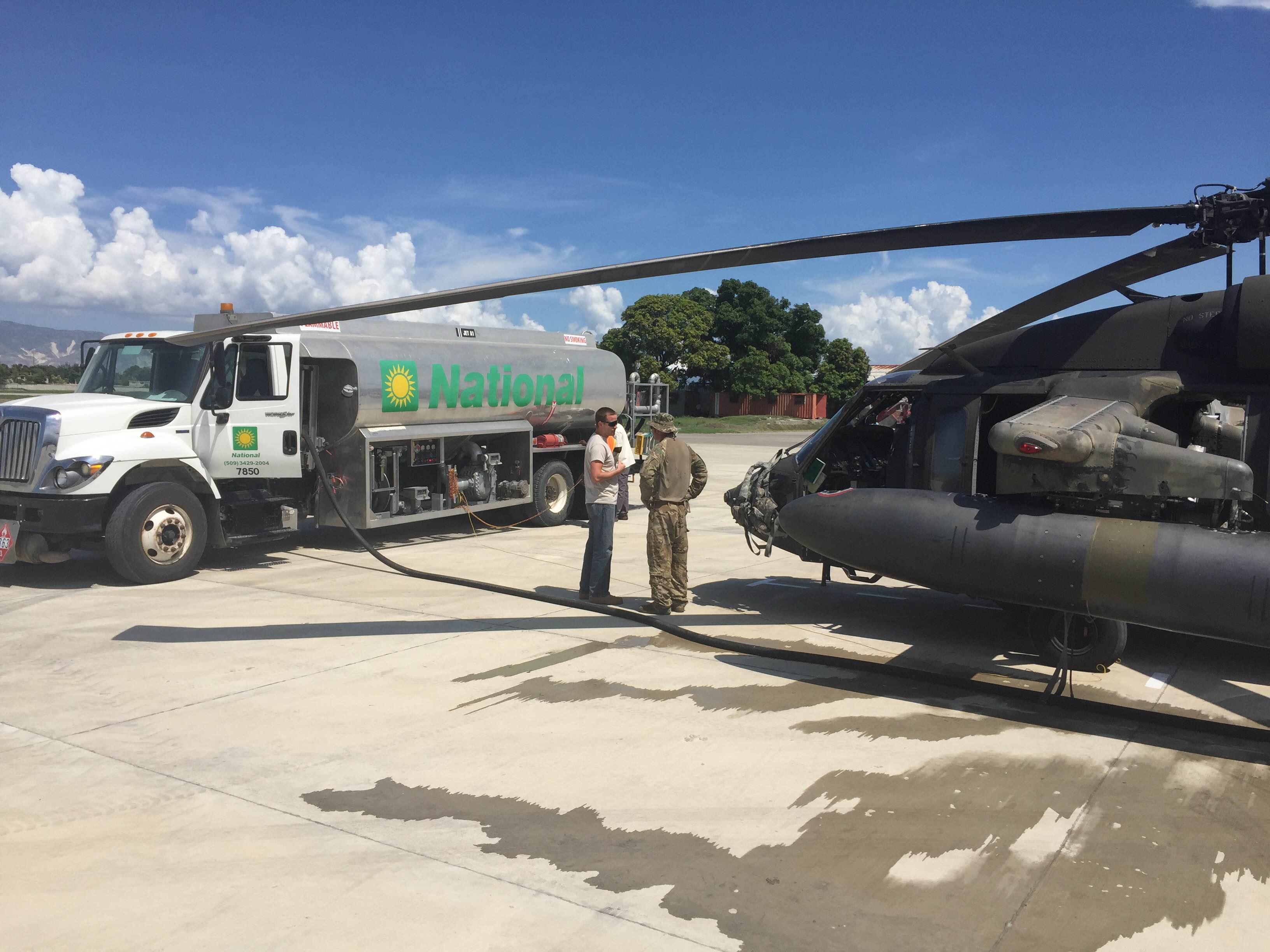 On landing pad, civilian, U.S. Army pilot in conversation between parked fuel truck and Army helicopter