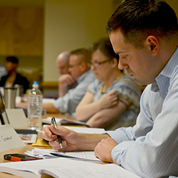 A man sitting at a long table with a woman and two other men while reading and filling out paperwork