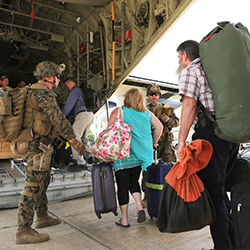 Men and women carrying luggage and being directed into an airplane by Marines.