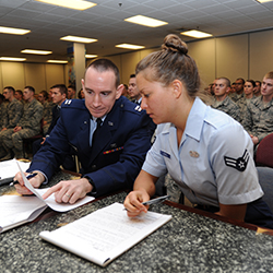 Two service members sitting at a table looking at papers with service members sitting in chairs behind them.