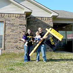 A husband and wife holding their children and sign in front of their house