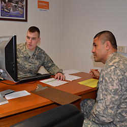 Two service members sitting at a desk looking at a computer monitor.