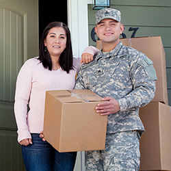 Service member holding a moving box and standing with his wife outside of their house