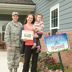 Female service member and a mother holding her daughter and a certificate in front of a house