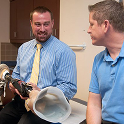 A man holding a prosthetic leg, sitting next to another man in a doctor’s office. 