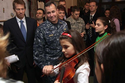 SKOPJE, Macedonia &mdash; Rear Adm. Andy Brown, European Command director of logistics, listens as a student at a local elementary school here plays the violin. Children from two schools in Macedonia showed their thanks Dec. 14 for refurbishments done on their schools. Individuals from the government of Macedonia, EUCOM, USAID and the U.S. Embassy donated money, time and supplies toward the school projects aimed toward bettering education in Macedonia. (Photo by Bekim Ajdini)