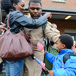 A service member reaching out to hug his wife and son, who is holding a flag in each hand.