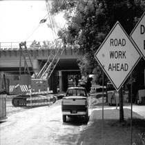 Picture depicts a bridge construction site within the Lansing, Michigan I-496 construction project.  A crane is visible near the bridge, along with a truck.  There is a Road Work Ahead sign in the foreground.