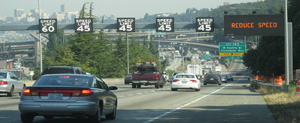 Photo. Dynamic lane control signs above a section of a Washington highway. Signage includes speed limits over 5 travel lanes and textual information in the far right lane.  Example of an ATM strategy in use – dynamic speed limits in Seattle, WA.