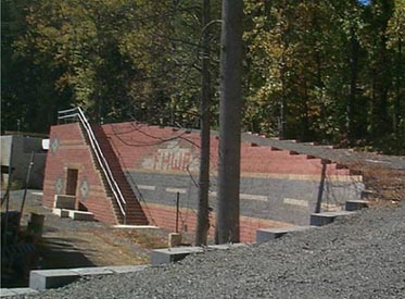 Photo. Long-range view of the FHWA prototype geosynthetic reinforced soil integrated bridge system. The facing modular block is decorative with FHWA inscribed and a road shown. A tunnel through the bridge and underneath a staircase is also depicted. 