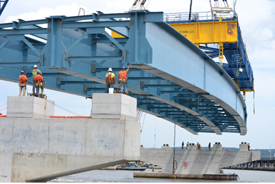 Title: Tappan Zee Bridge steel girder assembly. Photo Credit: New York State Thruway Authority - Description: A photograph of the new bridge under construction. A crane is lifting a large section of steel decking into place on concrete piers. Several construction workers and guiding the placement of the deck. 