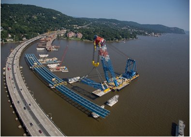 Title: Tappan Zee Bridge assembly. Photo Credit: New York State Thruway Authority. - Description: An ariel photograph of the new bridge construction adjacent to the existing bridge. A large crane is lifting a piece of steel decking into place on concrete piers. Several other sections are already in place. 