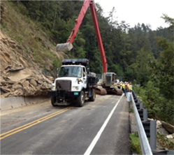 Photograph shows a rock slide on a road with an excavator in the background.