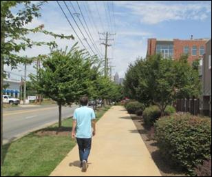 A young man walks down the newly redesigned sidewalk, which includes trees and grass separating him from the car lanes as well as additional landscaping.