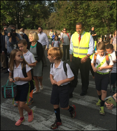 Secretary Foxx wears a reflective safety vest and walks across a street with a group of young children. 