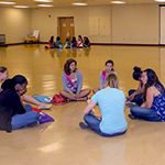 a group of young people sitting on a gym floor in a circle discussing topics of interest