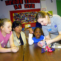 Three young girls watch their female teacher get crafts out onto a table 