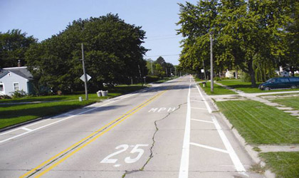 Photograph taken in Roland, Iowa, showing two parallel white lines on both sides of a curbed roadway with cross-hatch markings inside the two lines creating a shoulder area. A "25 MPH" is painted white in the center of the travel lane. There is a sidewalk offset from the right side of the roadway, and driveways can be seen on both sides of the road.