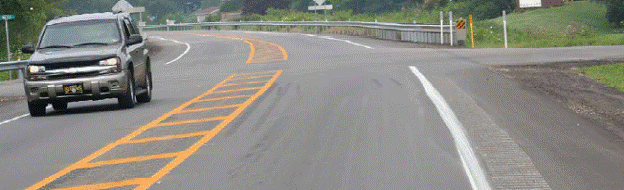 This figure shows an implementation of concept 1 at site 4 in Pennsylvania. The photo was taken from the major approach at an intersection of 2 two-lane, two-way roadways with stop-control on the minor approach. There are paved shoulders with a white edge line and yellow center line. Concept 1 includes the introduction of a painted yellow median island on the major road approaches. The painted median begins as a double yellow center line and gradually widens to a width of approximately 1.22 m (4 ft); the full median width is then continued to the intersection. Yellow cross-hatching is installed in the painted median. Rumble strips are also introduced on the outside shoulders and in the painted median on the major road approaches. A vehicle is shown travelling on the major road within the limits of concept 1.