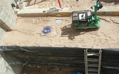 Top-down view showing into the outdoor geotechnical sand pits. A mechanically stabilized earth (MSE) shoring wall is on one side and a green lightweight vibratory compactor is shown on top of the sand.