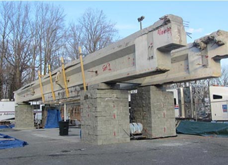 This photo shows four GRS piers with concrete masonry units (CMUs) on a concrete pad. Two large concrete I-beams are positioned on top of the GRS piers.  The beams have salt spray catch barriers attached to them.
