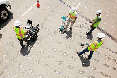 The photo is an overhead view of four workers in yellow safety vests and white helmets on a deck. The bridge deck is marked with white paint spots that form a grid pattern. The front fender and wheel of a white vehicle can be seen in the upper left corner of the photo, along with an orange traffic cone. The leftmost worker is pushing an instrument mounted on a three-wheeled cart. The instrument has a display screen. The worker in the middle of the image is holding a smaller computerized instrument which is in contact with the deck. The two workers on the right side of the image are holding probes.